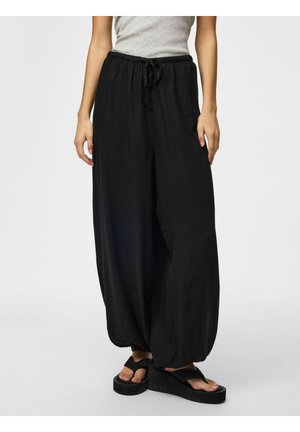 Donna che indossa pantaloni neri larghi con coulisse e sandali neri con zeppa, in piedi contro uno sfondo chiaro e uniforme.