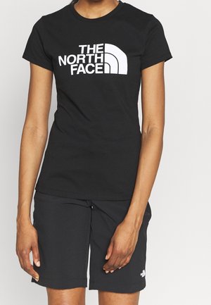 Persoon die een zwart T-shirt draagt met het witte "THE NORTH FACE"-logo en zwarte shorts met een klein bijpassend logo op het linkerbeen.