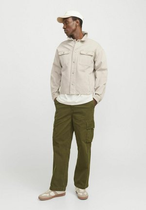 Hellbeige Denimjacke mit zwei Brusttaschen, kombiniert mit olivgrünen Cargo-Hosen mit mehreren Taschen und cremefarbenen Sneakers mit einem Muster.