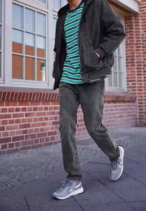 Person går på en fortov iført en sort jakke, grøn- og hvidstribet skjorte, grå jeans og grå sneakers nær en murstensbygning med vinduer.