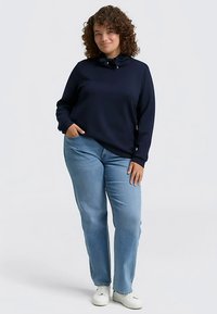 Marineblauwe sweatshirt met een hoge kraag en strikdetail, gecombineerd met lichtblauwe jeans met rechte pijpen en witte sneakers. Krullend haar zichtbaar.