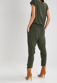 Une combinaison olive décontractée avec des manches courtes, une ceinture élastique et des jambes fuselées, associée à des bottines beiges.