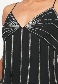 Czarna bluzka na ramiączkach, z ozdobami z srebrnych koralików ułożonymi w pionowe linie i wzory w formie wachlarzy. Miękka faktura materiału.