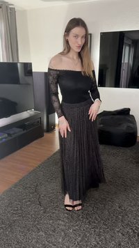 Zwarte off-shoulder top met kanten mouwen, gecombineerd met een lange, geplooide zwarte rok. Zwarte sandalen met open teen en bandjes.