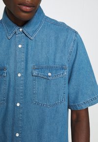 Světle modrá džínsová košile s krátkým rukávem, dvěma náprsními kapsami, kovovými druky a klasickým límečkem. Má jemnou texturu a viditelné švy.
