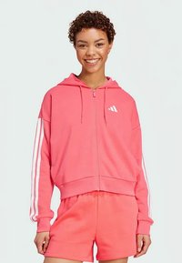 Rosa zip-up hoodie med vita ränder på ärmarna och logotyp. Tillverkad av mjukt tyg, med avslappnad passform och justerbar huva. Kombinerad med matchande shorts.