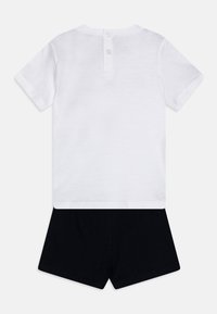 Kurzärmliges weißes Hemd mit Knopfverschluss am Rücken, kombiniert mit schwarzen Shorts. Hergestellt aus weichem Baumwollstoff mit glatter Textur.