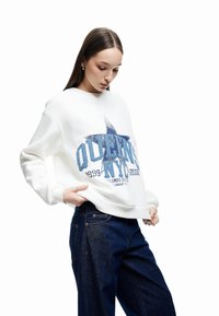 Sudadera blanca con un diseño gráfico azul que incluye estrellas y acentos de texto, combinada con jeans de pierna ancha de color azul oscuro.