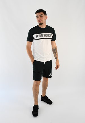 Conjunto deportivo en verde y blanco que incluye una camiseta de manga corta con la impresión "LE COQ SPORTIF", combinada con pantalones cortos oscuros y zapatillas negras.