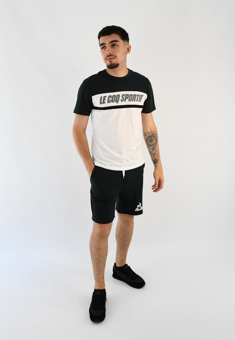 Conjunto deportivo en verde y blanco que incluye una camiseta de manga corta con la impresión "LE COQ SPORTIF", combinada con pantalones cortos oscuros y zapatillas negras.