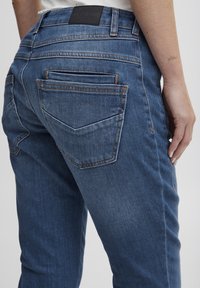 Tumed sinised denim teksad väikese pleekimisega. Omavad musta vööde sildikest, kahte tagakotti ja diskreetseid oranže õmblusi.