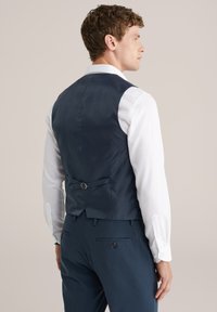 Mann in dunkelblauem Anzugweste und Hose, mit einem weißen Hemd, von hinten vor einem hellbeigen Hintergrund gezeigt.
