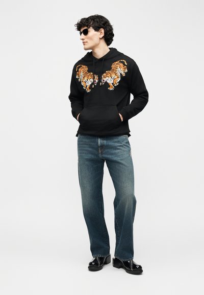 Νέος άνδρας που φοράει μαύρη μπλούζα με κουκούλα και σχέδια τίγρης, γυαλιά ηλίου, μπλε τζιν και μαύρες μπότες, στέκεται με τα χέρια στις τσέπες κοιτώντας προς τα αριστερά.