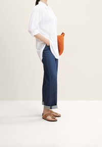 Weißes Hemd mit Knopfleiste, hochgekrempelte Ärmel, kombiniert mit blauen geraden Jeans und braunen Sandalen. Hält eine orange gesteppte Clutch.