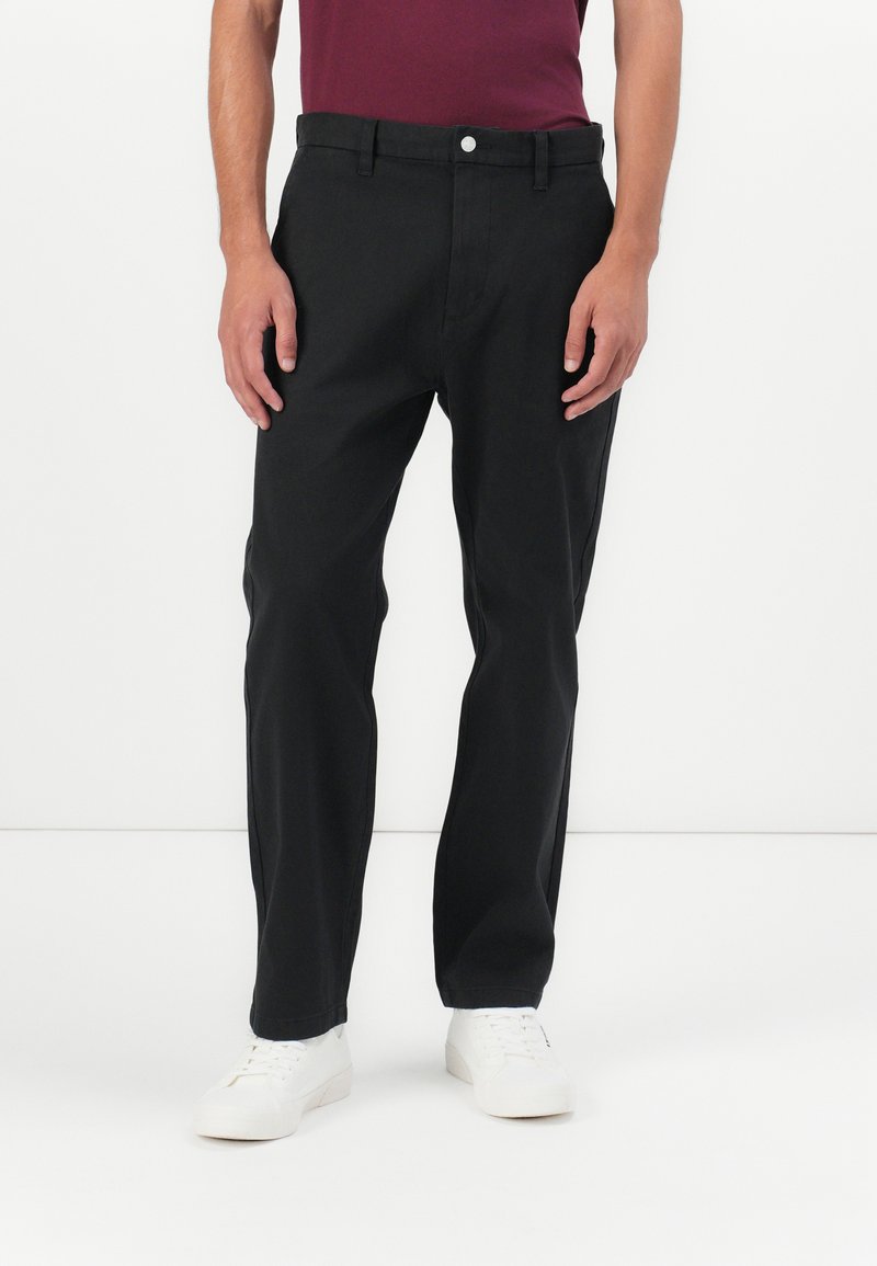 Schwarze Chino-Hosen mit geradem Bein, versehen mit einem Knopfverschluss, Gürtelschlaufen und Seitentaschen, kombiniert mit weißen Sneakers.