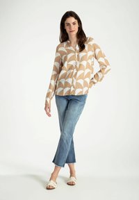 Beige Langarmhemd mit weißem Bogenmuster, Knopfverschluss, kombiniert mit hellblauen Jeans und beigen Slippern mit geflochtenem Design.