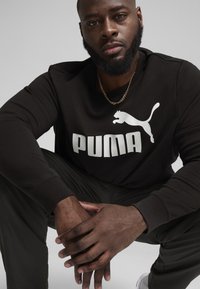 Čierna mikina s veľkým bielym logom "PUMA" a dizajnom skákania mačky. Model má na sebe tmavé nohavice a zlatý náhrdelník. Detailný záber.