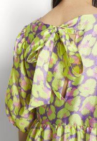 Vue de dos d'une robe brillante à motifs verts et violets avec des manches bouffantes et un grand nœud noué à l'encolure haute du dos.