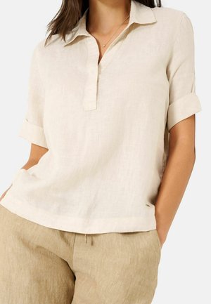 Femme portant une chemise en lin beige à manches courtes avec col et col en V, et un pantalon en lin marron clair, les mains dans les poches.