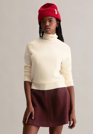 Cremfarbener Rollkragenpullover mit gerippten Bündchen und Saum, kombiniert mit einem kastanienbraunen A-Linien-Rock aus strukturiertem Stoff. Rote Strickmütze auf dem Kopf.