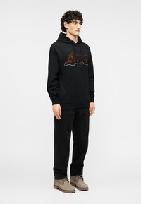 Μαύρη φούτερ με κουκούλα με σχέδιο γάτας από κόκκινα και λευκά στολίδια, μπροστινή τσέπη, φορεμένη με μαύρο παντελόνι και μπεζ μπότες.