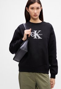 Čierna bavlnená mikina so strieborným logom Calvin Klein, doplnená malou čiernou koženou taškou s jedinečným tvarom a textúrovaným povrchom.