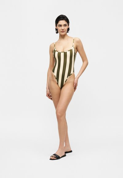 Γυναίκα που στέκεται, φοράει ολόσωμο μαγιό με κατακόρυφες ελαιόχρωμες και μπεζ ρίγες και μαύρα σαγιονάρα, σε λευκό φόντο.