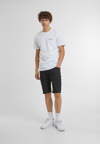 T-shirt bianca in cotone con logo blu, abbinata a pantaloni corti neri. Il modello indossa sneakers bianche e calze a lunghezza media. Design semplice, vestibilità comoda.