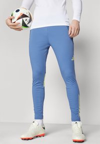 Blue athletic leggings made of stretchable material with green accents and Adidas logo; paired with white soccer cleats and holding a soccer ball.