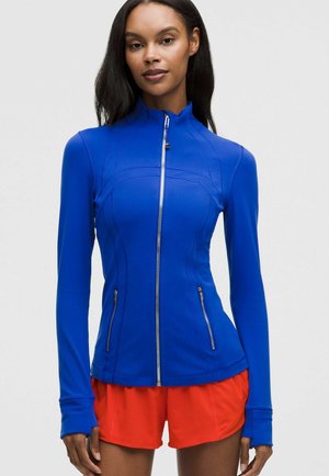 Vrouw draagt een nauwsluitend blauw jack met ritssluitingen en ritssluitingzakken en felrode sportshorts tegen een effen achtergrond.