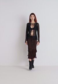 Haut en mesh noir à manches longues avec détails à nouer, associé à une jupe midi ajustée en franges marron. Le mannequin porte des bottines noires.