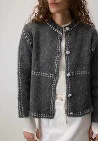 Ženska nosi temno sivo volneno cardigan jakno s srebrnimi gumbi in belim šiviljskim detajlom čez belo dolg rokav majico in bele hlače.