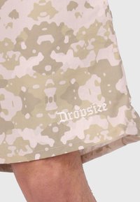 Tarnshorts in hellbeige und rosafarbenen Tönen, mit einem strukturierten Muster und dem gestickten "Dropsize"-Logo am Saum.