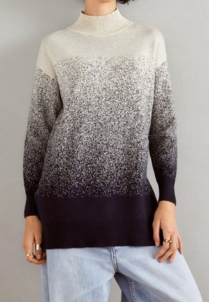 Persoon draagt een trui met een gevlekt verloop van lichtgrijs boven naar donkerblauw onder, gecombineerd met lichtblauwe spijkerbroek en ringen aan de vingers.