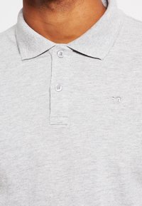 Man in een lichtgrijze gestructureerde polo met twee vastgemaakte knopen en een klein geborduurd logo op de borst.