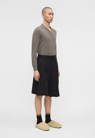 Άνδρας που στέκεται, φοράει μπλούζα πόλο μακρυμάνικη σε χρώμα ταουπέ, μαύρη πλισέ φούστα μέχρι το γόνατο, μαύρες κάλτσες και μπεζ χνουδωτές παντόφλες σε λευκό φόντο.