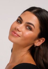 Nærbilde av en person med middels hudtone og mørkt, rett hår, iført gullbøylerøreringer og svart topp. Klar hud og skinnende lepper.