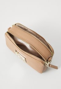 Piccolo borsa a tracolla in pelle color beige con due scomparti frontali aperti con zip dorate e un emblema decorativo dorato sul davanti.