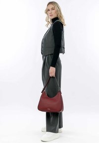 Gilet grigio con bottoni sopra un dolcevita nero, abbinato a pantaloni grigi a gamba larga. Tiene una borsa hobo in pelle rossa con una forma curvata.