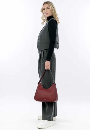 Gilet grigio con bottoni sopra un dolcevita nero, abbinato a pantaloni grigi a gamba larga. Tiene una borsa hobo in pelle rossa con una forma curvata.