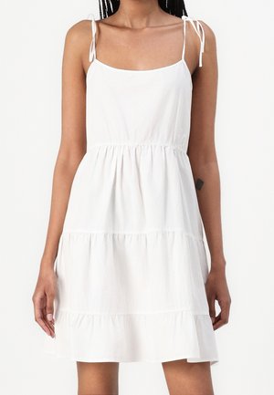 Vrouw in een mouwloze witte tiered jurk met spaghetti bandjes, staande tegen een effen witte achtergrond.