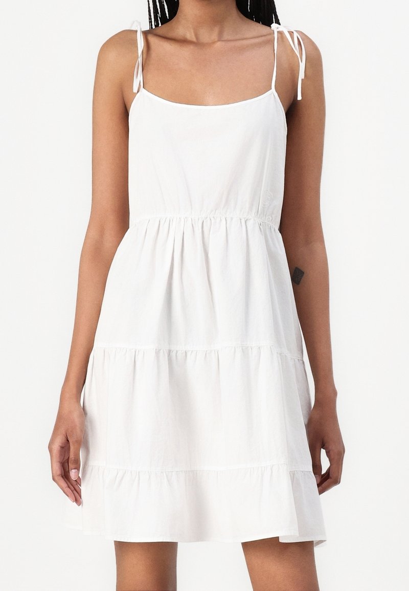 Kobieta w białej sukience bez rękawów z warstwami i cienkimi ramiączkami, stojąca na tle jednolitej białej ściany.