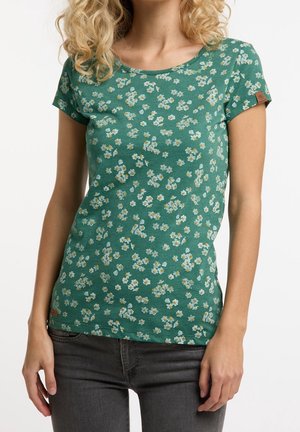 Mujer con camisa verde de manga corta con pequeño estampado floral blanco y amarillo y vaqueros gris oscuro, de pie con postura relajada.