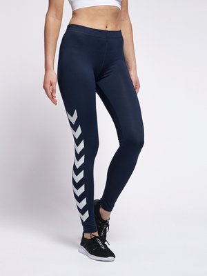 Vrouw in marineblauwe leggings met witte chevronstrepen op het linkerbeen, zwarte sportschoenen en een witte sportbeha, staand tegen een witte achtergrond.