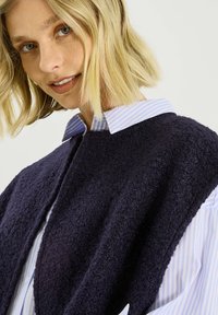 Chaleco de forro polar azul marino con superficie texturizada y frente abierto, superpuesto sobre una camisa de botones a rayas azul claro y blanca.