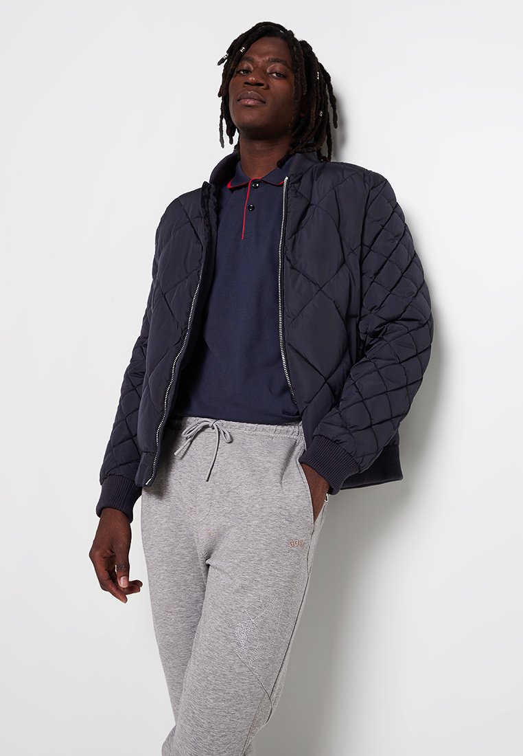 Veste bomber matelassée marine avec fermeture éclair et poignets côtelés, associée à un polo marine et à un pantalon de jogging gris, doté d'un cordon de serrage ajustable.