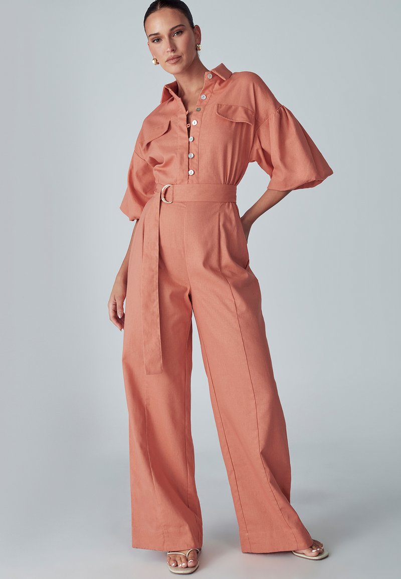 Mujer con un mono coral de pierna ancha y cinturón, con mangas cortas y frente abotonado, sandalias de tacón beige y pendientes de aro dorados.