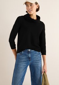 Zwart coltrui met driekwartmouwen, gecombineerd met lichtblauwe rechtlopende jeans. De jeans hebben een textured patroon en zijstrepen.