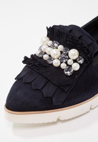 Mocassin en daim noir avec semelle blanche, décoré de cuir à franges et orné de perles blanches regroupées et de strass transparents sur le dessus.