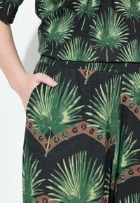 Bloemenprint broek heeft groene palmbladpatronen op een zwarte achtergrond, met een elastische tailleband en zijzakken voor functionaliteit.